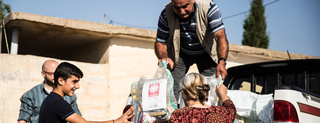 Humanitäre Hilfe in Syrien Ein Mann steht auf der Ladefläche eines kleinen LKWs und gibt Säcke mit Gewand an Menschen unten weiter. Auf den Säcken ist das Logo von Caritas Syrien.