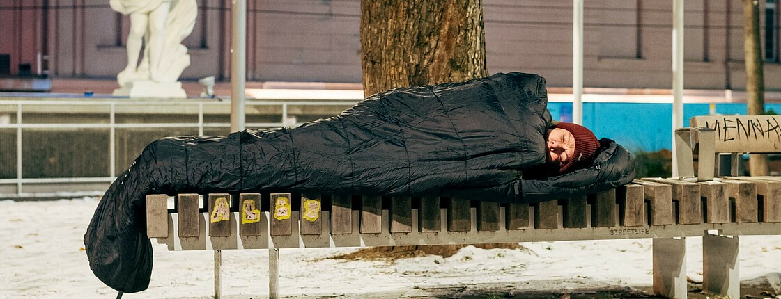 Eine Person schläft auf einer Parkbank, sie ist in einen Schlafsack gewickelt. Es ist winterlich.