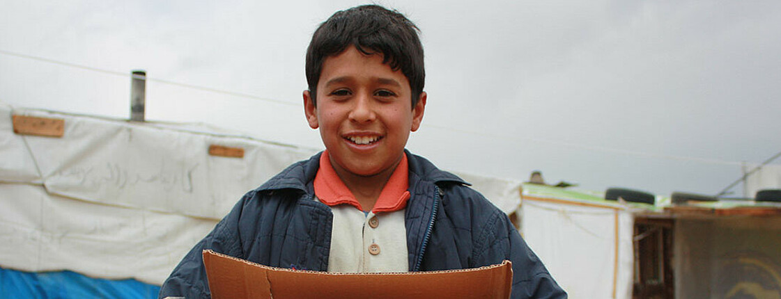 Ein Flüchtlingskind befindet sich im Libanon in einem Zeltlager. Er hält eine große Schachtel in den Händen. 