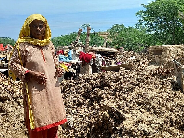 Eine Frau steht inmitten von Trümmern und Schlamm, die Reste eines zerstörten Hauses sind zu erkennen.