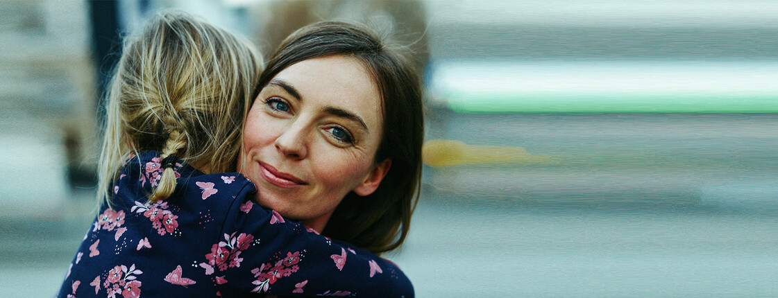 Eine Frau in einer geblümten Bluse umarmt ein Kind und lächelt warm, während der Hintergrund verschwommen bleibt