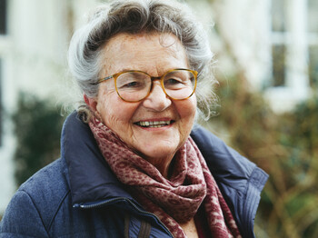 Fachstelle für Pflegende Angehörige Portrait einer Seniorin mit Brille, die einen roten Schal und einen blauen Wintermantel trägt.