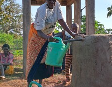 Eine ältere südsudanesische Frau füllt eine Giesskanne an einem Wasserhahn. 