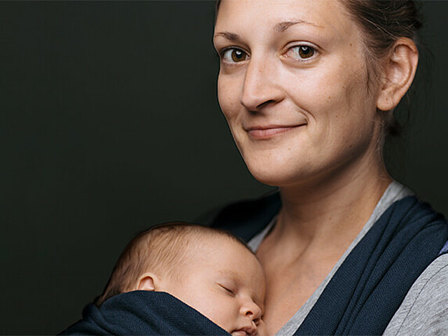 Eine junge Frau lächelt in die Kamera, sie trägt ein Baby im Tragetuch vor sich. Der Hintergrund ist schwarz.