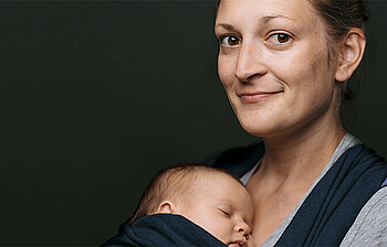 Eine junge Frau lächelt in die Kamera, sie trägt ein Baby im Tragetuch vor sich. Der Hintergrund ist schwarz.