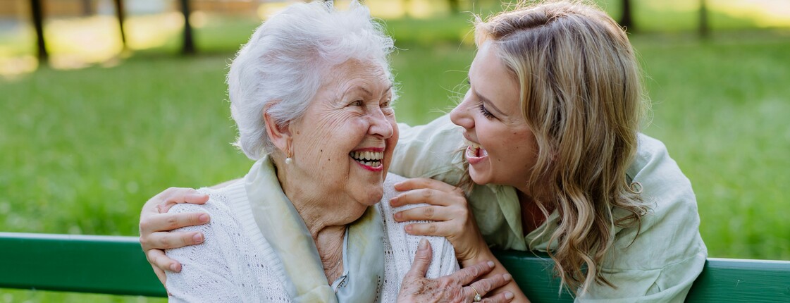 In einem Park sitzt eine Seniorin auf einer grünen Bank. Hinter ihr steht eine junge Frau, die ihre Hände auf die Schultern der Seniorin gelegt hat. Beide schauen sich lachend an.