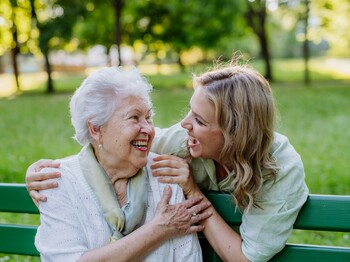Demenz-Servicezentrum In einem Park sitzt eine Seniorin auf einer grünen Bank. Hinter ihr steht eine junge Frau, die ihre Hände auf die Schultern der Seniorin gelegt hat. Beide schauen sich lachend an.