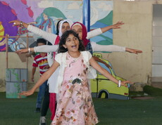 Mädchen mit ausgestreckten Armen tanzen auf grünem Boden vor farbenfroher Wand im Bekaa-Tal, Libanon