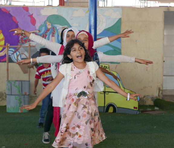 Mädchen mit ausgestreckten Armen tanzen auf grünem Boden vor farbenfroher Wand im Bekaa-Tal, Libanon