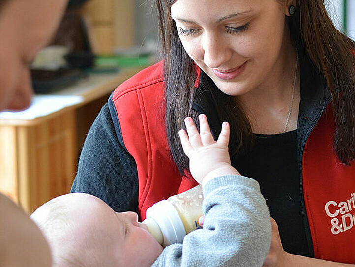 Frau mit Caritas-Jacke füttert ein Baby mit einem Fläschchen.