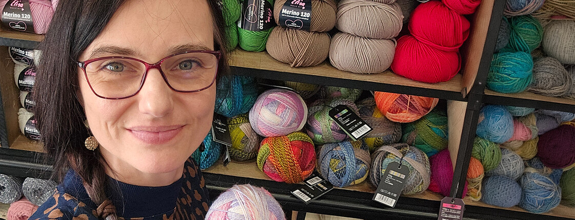 Person hält Wollknäuel in Stoffgeschäft vor Regalen mit Wolle beim Mützchen-Stricken für Caritas und Innocent.