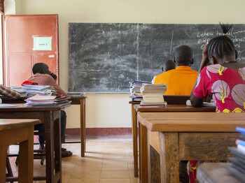Drei Schüler sitzen von hinten an Holzschulbänken in einem einfachen Klassenzimmer im Südsudan, auf den Tischen liegen Schulbücher und Hefte.