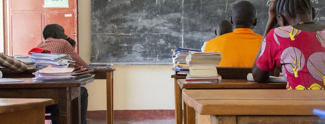 Drei Schüler sitzen von hinten an Holzschulbänken in einem einfachen Klassenzimmer im Südsudan, auf den Tischen liegen Schulbücher und Hefte.