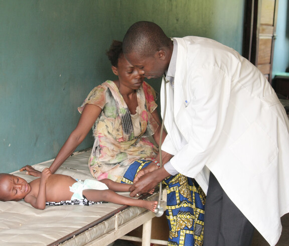 Ein Arzt untersucht ein krankes Kleinkind, das weinend auf einer Liege neben seiner Mutter liegt