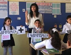Eine Lehrerin hält in einem Klassenzimmer Unterricht. Neben ihr stehen Schülerinnen, die Schilder mit armenischen Wörtern halten..