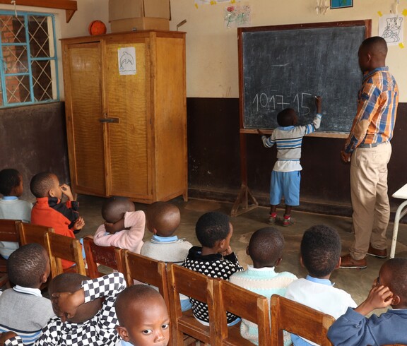 Ein kleines Kind schreibt unter Anleitung eines Lehrers eine Rechenaufgabe an eine Kreidetafel; davor sitzen zahlreiche Kinder auf Holzstühlen und beobachten aufmerksam – Grundschulunterricht in einer von der Caritas unterstützten Schule in Burundi.