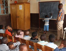 Ein kleines Kind schreibt unter Anleitung eines Lehrers eine Rechenaufgabe an eine Kreidetafel; davor sitzen zahlreiche Kinder auf Holzstühlen und beobachten aufmerksam – Grundschulunterricht in einer von der Caritas unterstützten Schule in Burundi.
