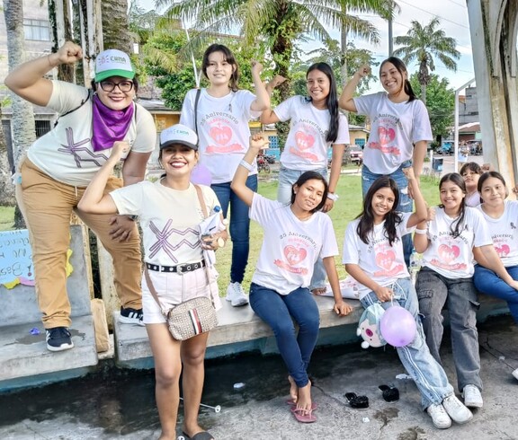 Eine Gruppe von Mädchen und jungen Frauen in weißen Jubiläums-T-Shirts posiert lachend und mit erhobenen Fäusten auf einem Platz in Iquitos; im Hintergrund sind weitere Teilnehmerinnen einer Veranstaltung zu sehen