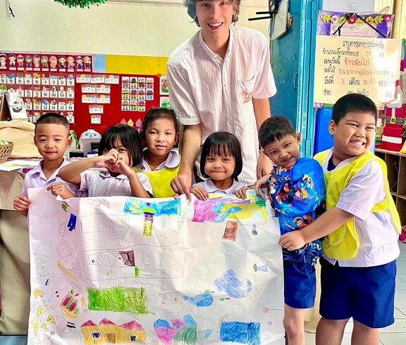 Ein junger Erwachsener steht mit mehreren Kindern in einem Klassenzimmer; gemeinsam halten sie ein großes, bemaltes Papier.