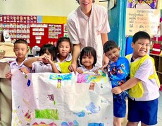 Ein junger Erwachsener steht mit mehreren Kindern in einem Klassenzimmer; gemeinsam halten sie ein großes, bemaltes Papier.