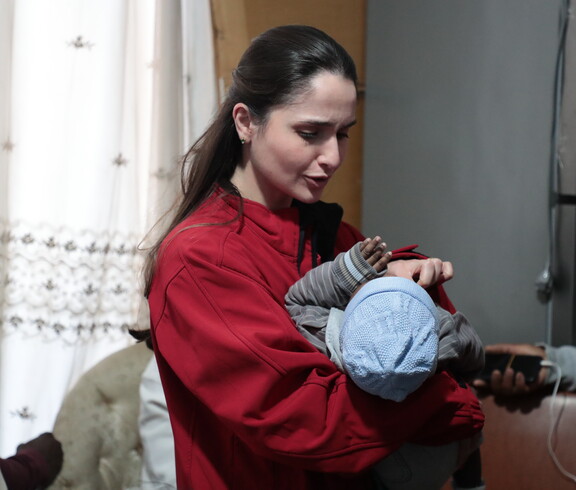 Frau in roter Jacke hält ein Baby im Arm und spricht mit anderen Menschen.