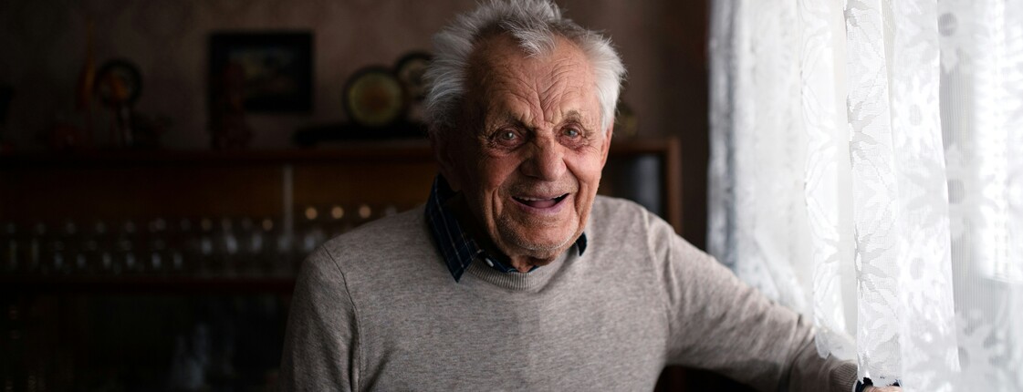 Ein lachender Mann im hohen Alter steht an einem Licht durchfluteten Fenster.