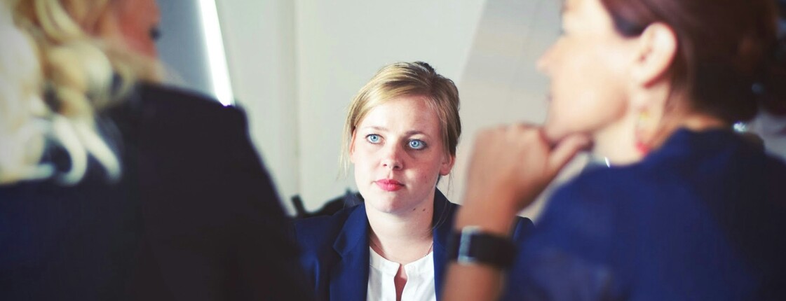 Drei Frauen sitzen in einem Gespräch, im Vordergrund ist eine Person unscharf, im Hintergrund eine Frau mit blondem Haar im Fokus.