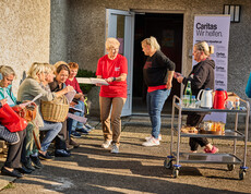 Mehrere Frauen sitzen auf einer Bank und besprechen Dokumente, während eine Caritas-Helferin ihnen Unterlagen überreicht. Im Hintergrund steht ein Tisch mit Getränken und Lebensmitteln sowie ein Caritas-Banner mit der Aufschrift „Caritas Wir helfen“.