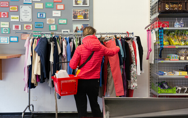 Eine Kundin im carla Shop in Tirol stöbert durch einen Kleiderständer mit Secondhand-Kleidung, während sie einen roten Einkaufskorb trägt.