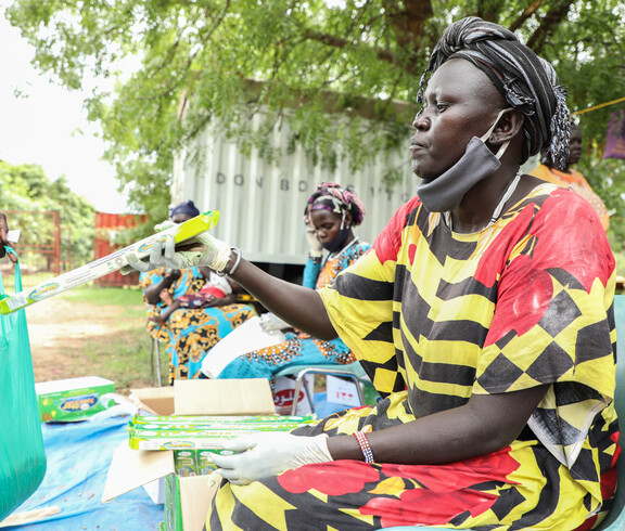 Eine Frau mit Mund-Nasen-Schutz nimmt ein Päckchen Öl entgegen, im Hintergrund warten weitere Frauen bei einer Hilfsgüterverteilung im Freien in Südsudan.