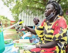 Eine Frau mit Mund-Nasen-Schutz nimmt ein Päckchen Öl entgegen, im Hintergrund warten weitere Frauen bei einer Hilfsgüterverteilung im Freien in Südsudan.