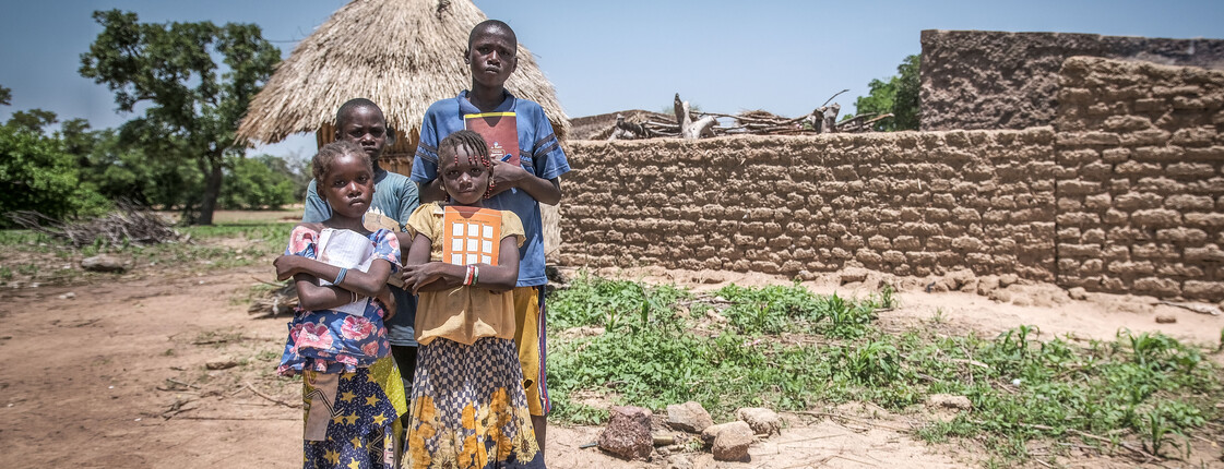 Vier Schulkinder stehen mit Schulheften unter dem Arm vor einem kleinen Haus in Burkina Faso. 