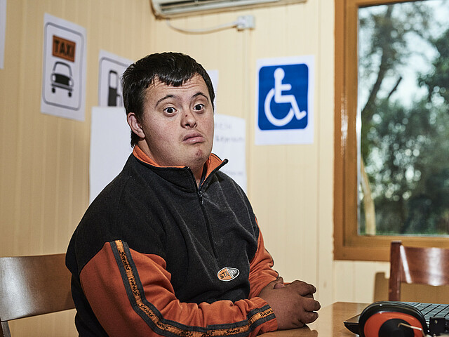 Der Oberkörper eines jungen Mannes ist zu sehen, im Hintergrund ein Zeichen an der Wand mit einem Rollstuhl