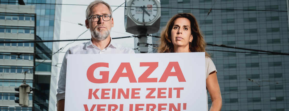 Alexander Bodmann, Vizepräsident der Caritas Österreich und Laura Leyser, Geschäftsführerin von Ärzte ohne Grenzen Österreich halten ein Schild mit dem Schriftzug "GAZA keine Zeit verlieren". Im Hintergrund eine Uhr.