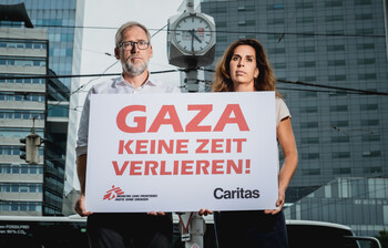 Alexander Bodmann, Vizepräsident der Caritas Österreich und Laura Leyser, Geschäftsführerin von Ärzte ohne Grenzen Österreich halten ein Schild mit dem Schriftzug "GAZA keine Zeit verlieren". Im Hintergrund eine Uhr.