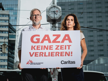 Alexander Bodmann, Vizepräsident der Caritas Österreich und Laura Leyser, Geschäftsführerin von Ärzte ohne Grenzen Österreich halten ein Schild mit dem Schriftzug "GAZA keine Zeit verlieren". Im Hintergrund eine Uhr.