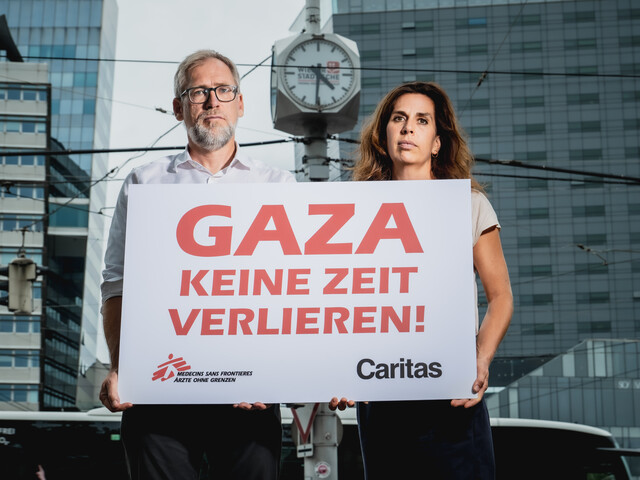Alexander Bodmann, Vizepräsident der Caritas Österreich und Laura Leyser, Geschäftsführerin von Ärzte ohne Grenzen Österreich halten ein Schild mit dem Schriftzug "GAZA keine Zeit verlieren". Im Hintergrund eine Uhr.