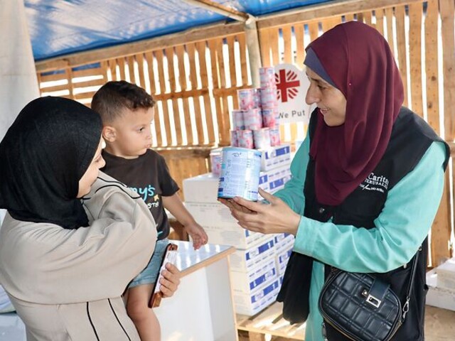 Eine Mitarbeiterin der Caritas Jerusalem zeigt einer Frau mit kleinem Kind eine Dose Babynahrung. Im Hintergrund sind Hilfspakete und das Caritas-Logo zu sehen.