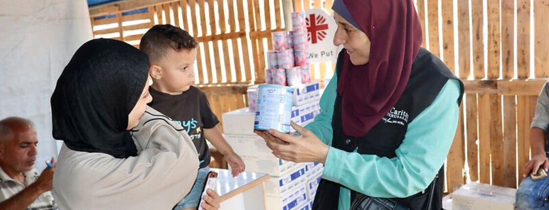 Eine Mitarbeiterin der Caritas Jerusalem zeigt einer Frau mit kleinem Kind eine Dose Babynahrung. Im Hintergrund sind Hilfspakete und das Caritas-Logo zu sehen.