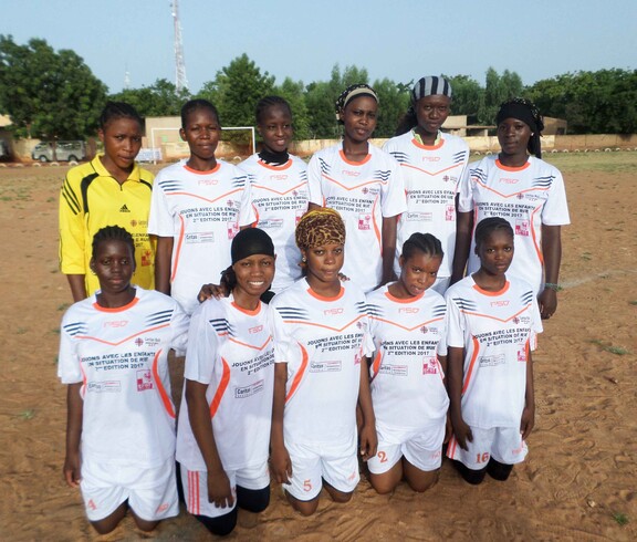 Eine Mädchenfußballmannschaft in Mali posiert gemeinsam für ein Foto. Sie tragen weiße Trikots und stehen auf einem Sportplatz.