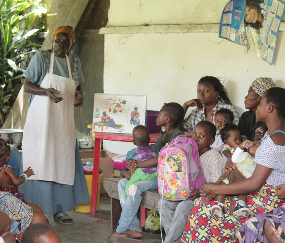 Eine Krankenschwester erklärt Eltern mit Hilfe von Anschauungsmaterial, wie sie ihre Kinder ausgewogen und nahrhaft ernähren können. Die Eltern hören aufmerksam zu und halten ihre Kinder im Arm 