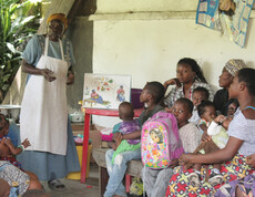 Eine Krankenschwester erklärt Eltern mit Hilfe von Anschauungsmaterial, wie sie ihre Kinder ausgewogen und nahrhaft ernähren können. Die Eltern hören aufmerksam zu und halten ihre Kinder im Arm 