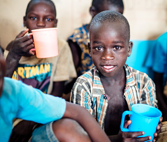 Ein Kind mit einer Zahnlücke vorne, lächelt in die Kamera, während er einen blauen Becher in der Hand hält. Um ihn herum sind seine Klassenkameraden, die etwas aus dem Bechern trinken.