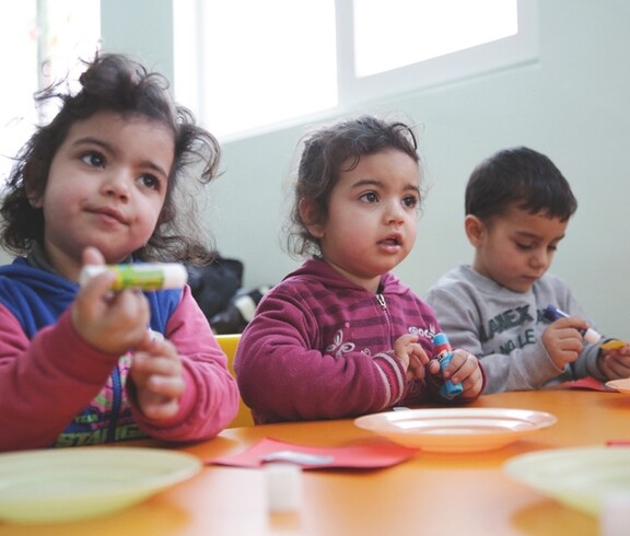 Drei kleine Kinder sitzen an einem Tisch und halten bunte Bastel- oder Klebestifte in den Händen.