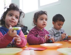 Drei kleine Kinder sitzen an einem Tisch und halten bunte Bastel- oder Klebestifte in den Händen.