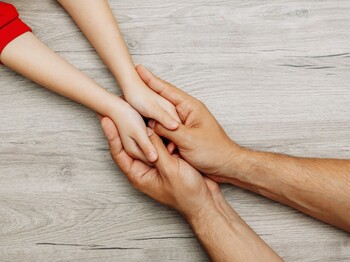 Man sieht zwei große Hände, die die Hände von einem Kind halten. 