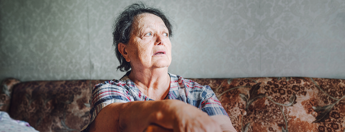 Eine ältere Dame sitzt auf der Couch, sie stützt sich auf ihren Stock und sieht aus dem Fenster.