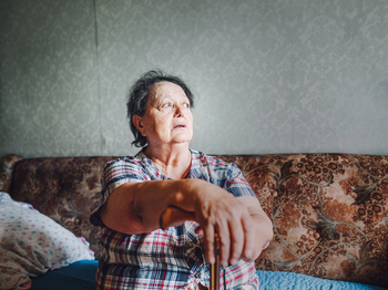 Eine ältere Dame sitzt auf der Couch, sie stützt sich auf ihren Stock und sieht aus dem Fenster.