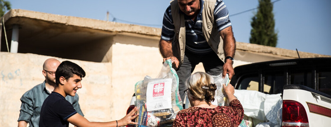 Ein Mann steht auf der Ladefläche eines kleinen LKWs und gibt Säcke mit Gewand an Menschen unten weiter. Auf den Säcken ist das Logo von Caritas Syrien.