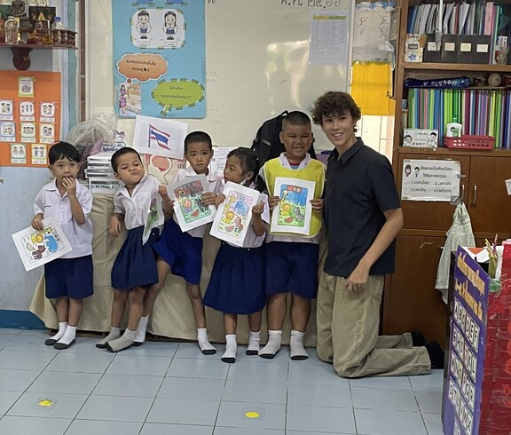Mehrere Kinder stehen in einem Klassenzimmer und halten bemalte Blätter hoch; ein junger Erwachsener kniet neben ihnen.